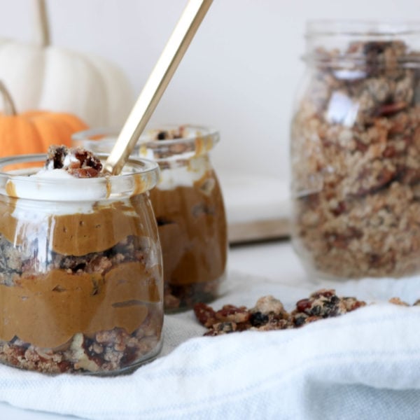 Two jars filled with Deconstructed Pumpkin Pie on a counter with a white kitchen towel and a jar of granola and pumpkins in the background. Ingredients include pumpkin, nut milk, prunes, maple syrup, vanilla.
