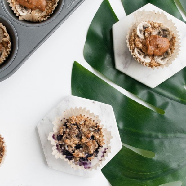 Two muffins on marble coasters on top of a green leaf on a white surface. Ingredients include banana, egg, coconut oil, rolled oats, almond milk.