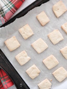 Short bread cookie squares on parchment paper placed on a baking sheet with a red Christmas kitchen towel.