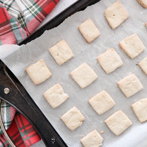Short bread cookie squares on parchment paper placed on a baking sheet with a red Christmas kitchen towel.