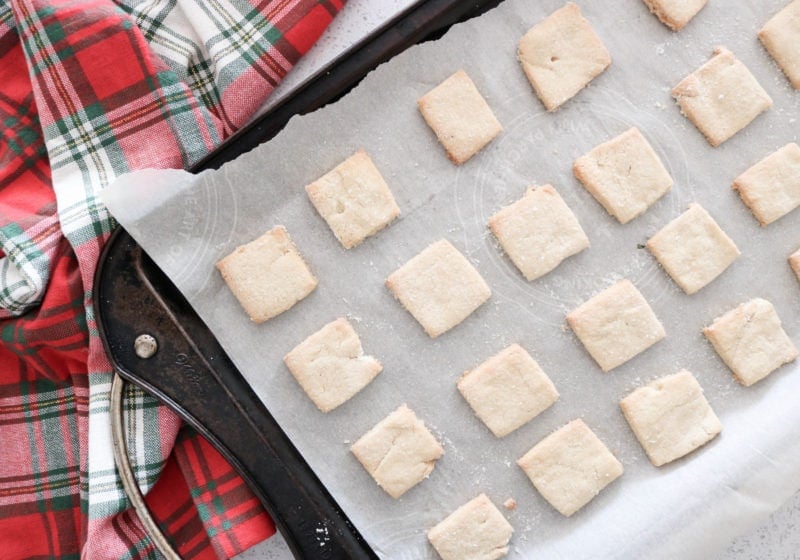 Short bread cookie squares on parchment paper placed on a baking sheet with a red Christmas kitchen towel.