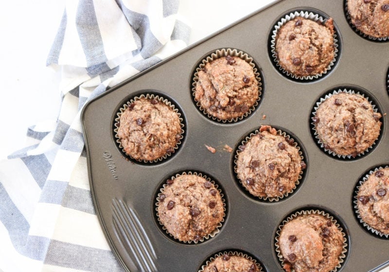 A muffin tin filled with baked muffins with a kitchen towel placed beside them. Ingredients include chocolate chips, banana, zucchini.