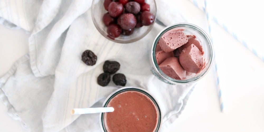 A photo of a dark chocolate cherry prune smoothie on a white food photography board with white kitchen towels and some prunes placed around it.