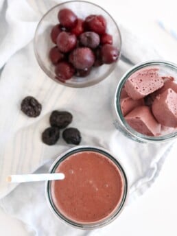 A photo of a dark chocolate cherry prune smoothie on a white food photography board with white kitchen towels and some prunes placed around it.