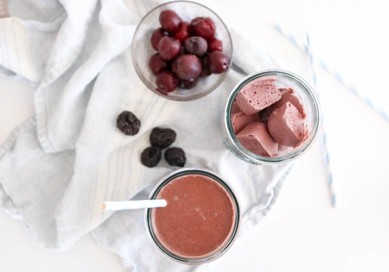 A photo of a dark chocolate cherry prune smoothie on a white food photography board with white kitchen towels and some prunes placed around it.