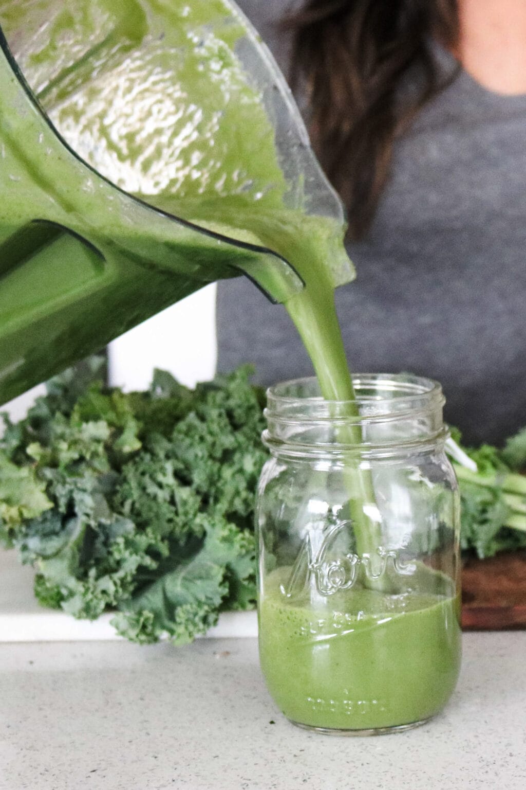 A vitamix blender is pouring green juice into a mason jar. There is a cutting board in the background with curly kale on it. 