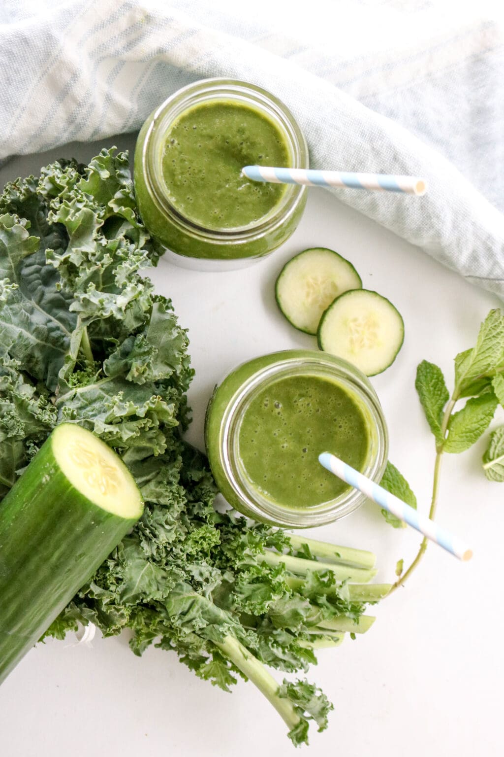 An overhead shot of two mason jars with straws filled with green juice. There is kale and cucumber to the left of the glasses. 