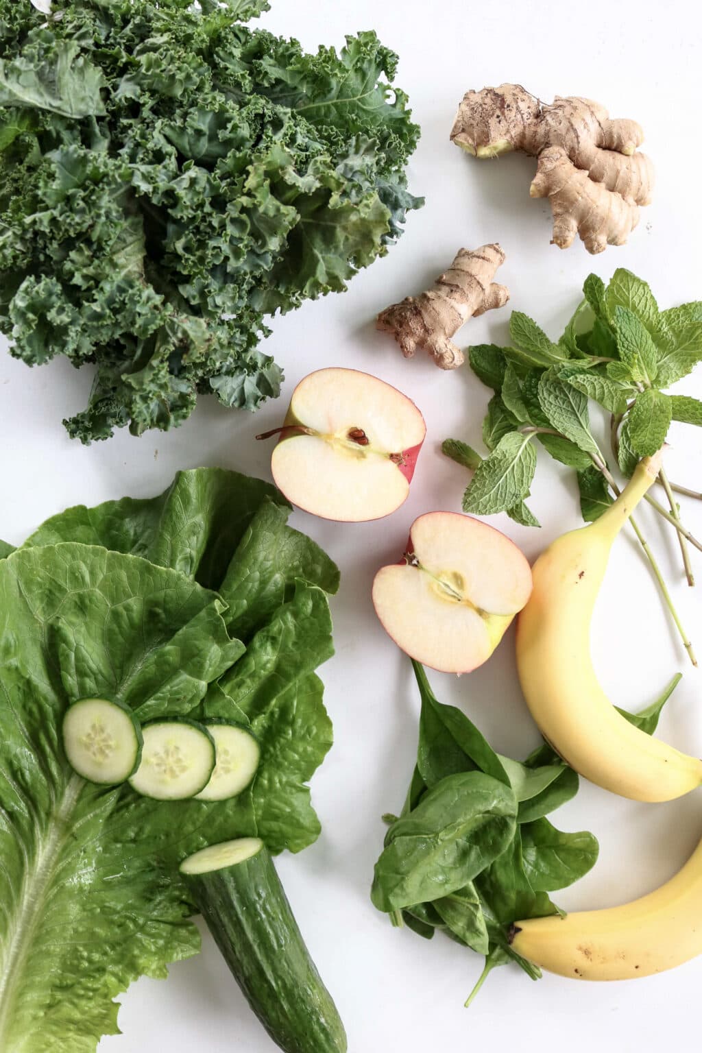 An overhead shot of ingredients for the green juice. There is curly green kale, two thumb-sized pieces of ginger, an apple cut in half, mint, a banana, romaine lettuce, and cucumber