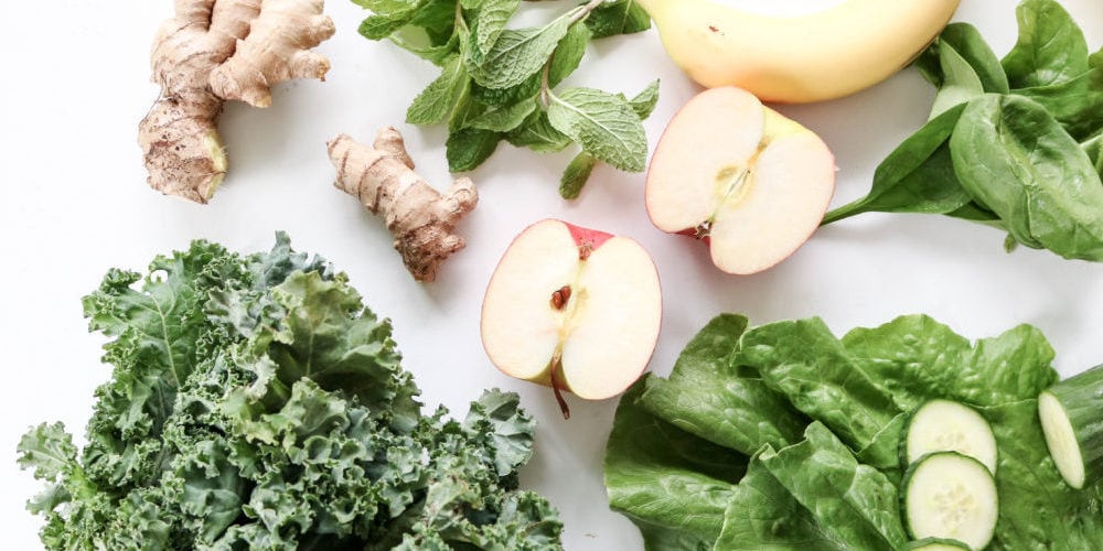 Ingredients for a green juice which supports good digestion laid out on a white cutting board. Ingredients include kale romaine apples banana ginger and mint