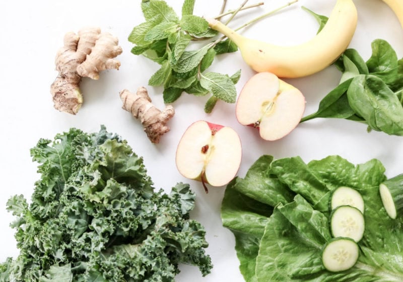 Ingredients for a green juice which supports good digestion laid out on a white cutting board. Ingredients include kale romaine apples banana ginger and mint