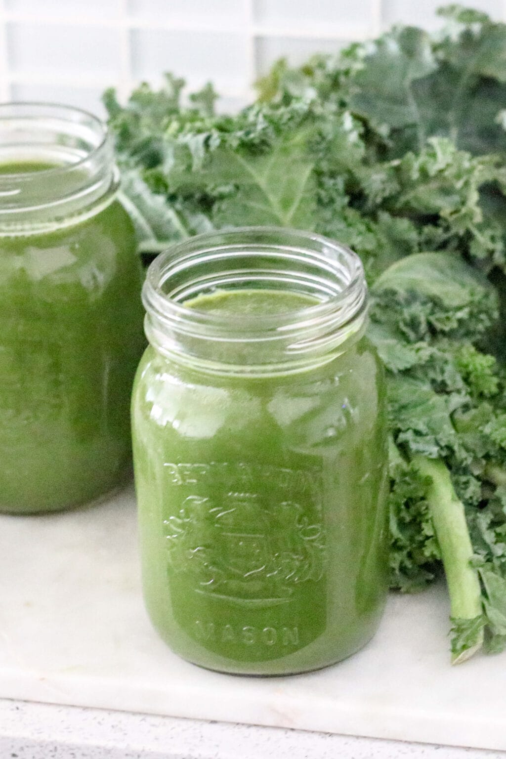 Two mason jars filled with green juice are sitting on a counter. Behind the cups is curly kale. 