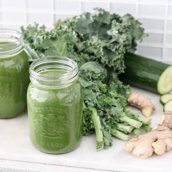 Two glass jars filled with green juice on the counter with vegetables in the background. Ingredients include kale, cucumber, ginger.