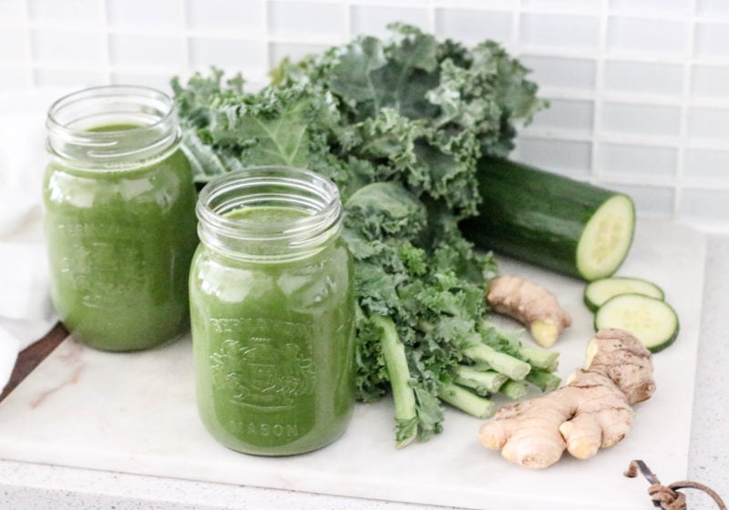 Two glass jars filled with green juice on the counter with vegetables in the background. Ingredients include kale, cucumber, ginger.