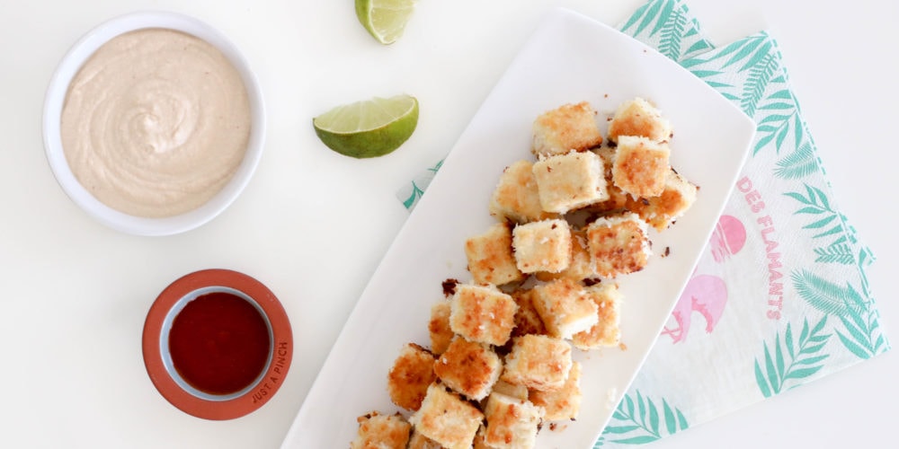 Crispy tofu bites placed on a rectangular white serving dish surrounded by sauce and lime wedges. Ingredients include tofu, coconut, bread crumbs.