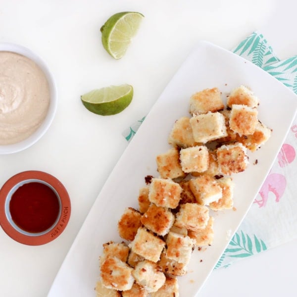 Crispy tofu bites placed on a rectangular white serving dish surrounded by sauce and lime wedges. Ingredients include tofu, coconut, bread crumbs.