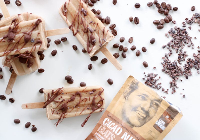 Three Coconut Cream Coffee Popsicle's on a white surface with coffee beans spread around them. Ingredients include coffee, coconut cream, chocolate chips, cacao nibs.