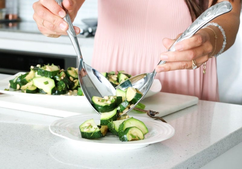 Registered Dietitian tossing zucchini with two big silver serving spoons.