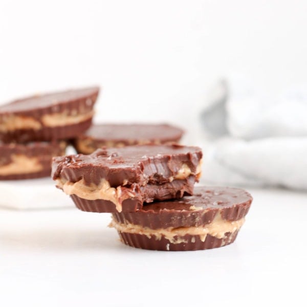 Chocolate peanut butter cups placed on a white surface with a white kitchen towel in the background. Ingredients include chocolate, coconut oil, peanut butter.