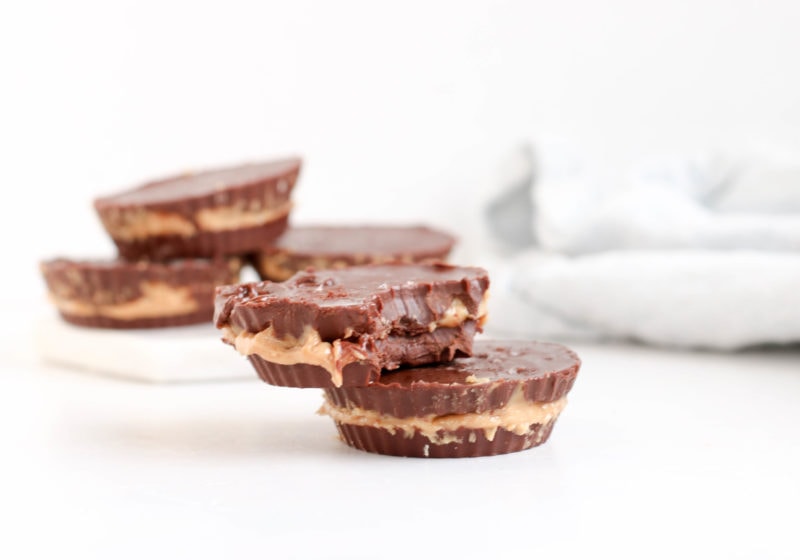 Chocolate peanut butter cups placed on a white surface with a white kitchen towel in the background. Ingredients include chocolate, coconut oil, peanut butter.