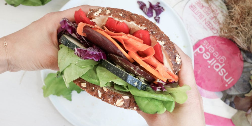Registered Dietitian Lindsay Pleskot holding a sandwich over top of a white round place. Ingredients include bread, greens, hummus, avocado, carrots and zucchini.