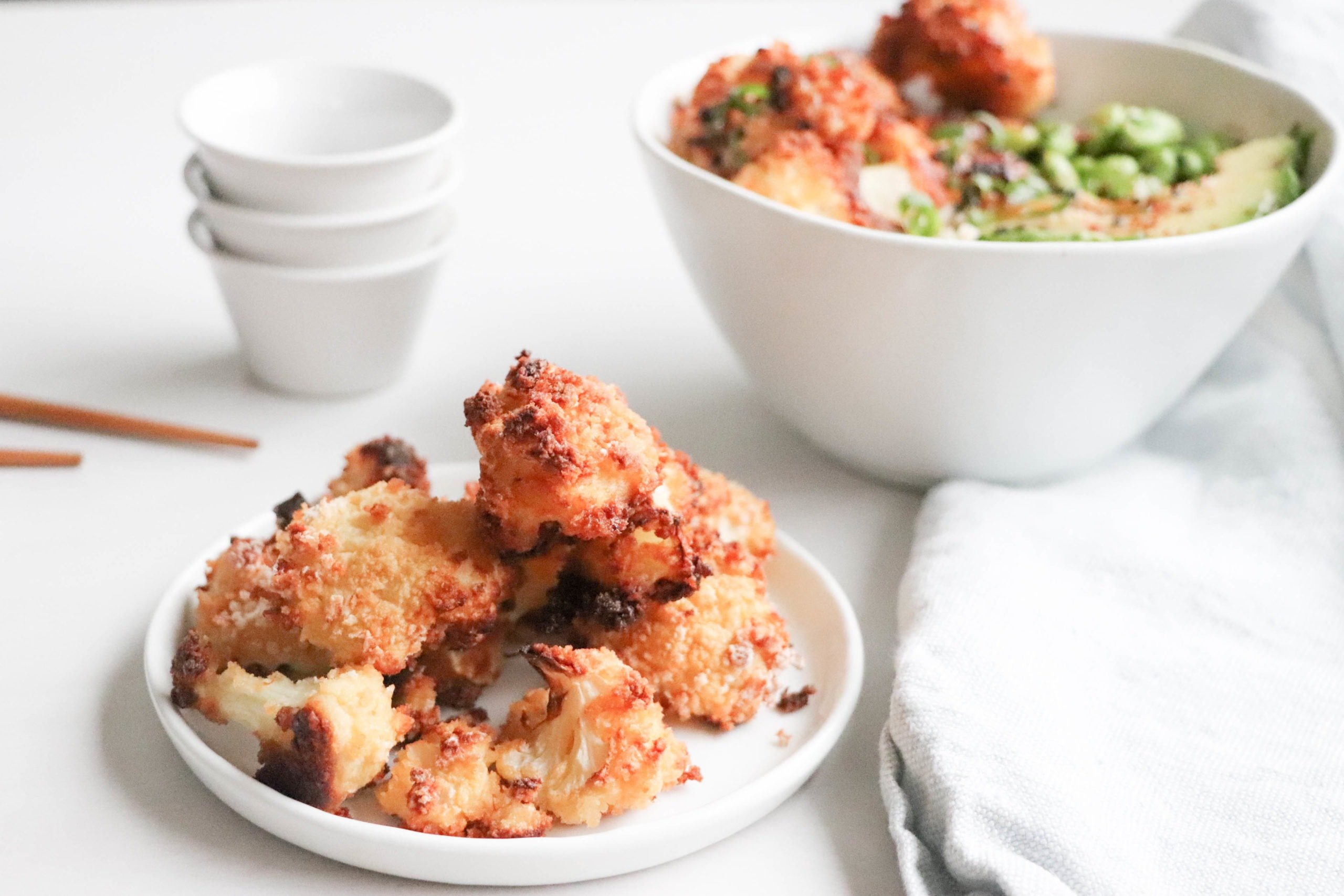 Crispy baked cauliflower on a white plate over a white surface with a white kitchen towel and stack of white small bowls.