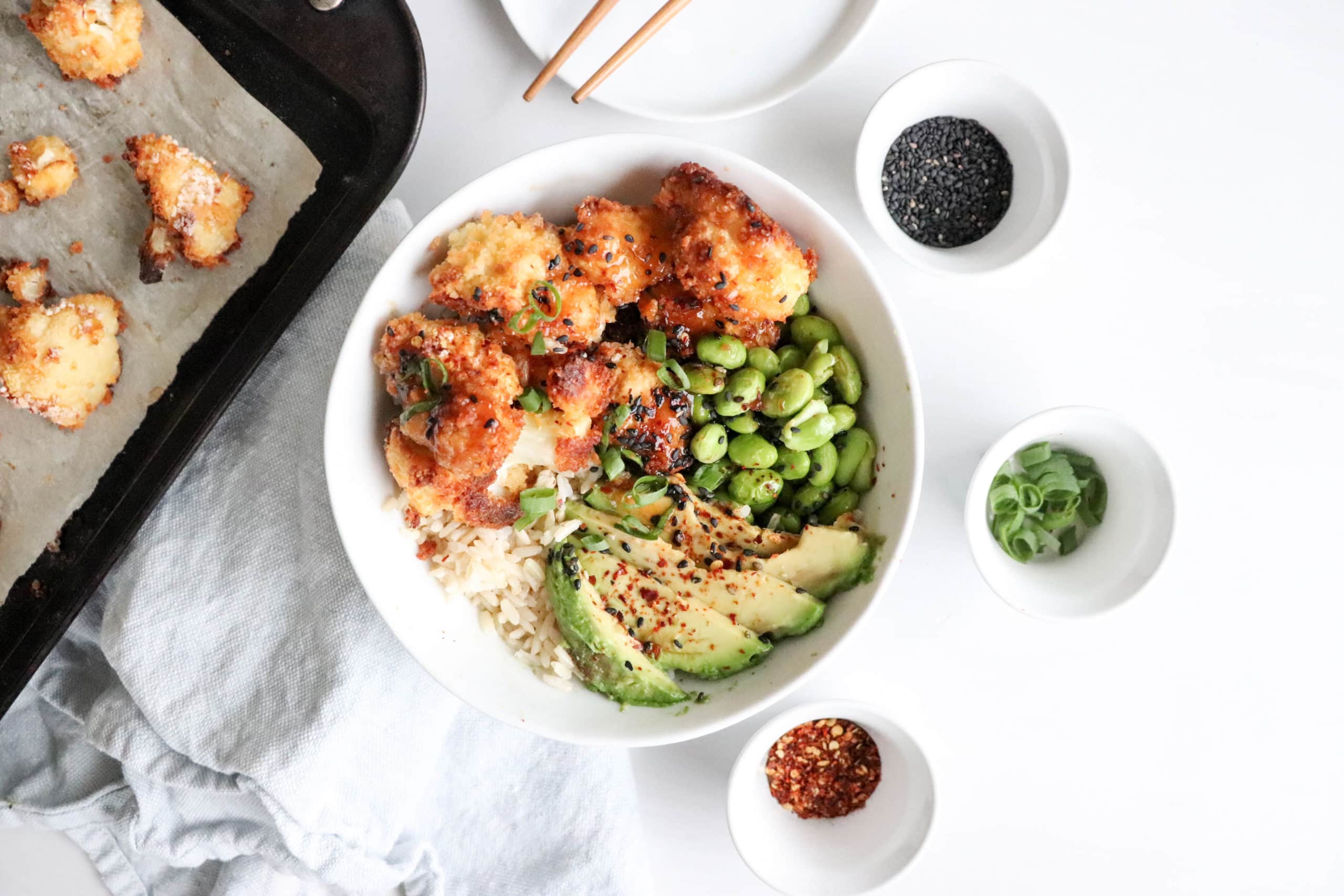 Crispy baked cauliflower bowl on a white surface with a blue kitchen towel underneath it. Ingredients include: rice, avocado, edamame, crispy baked cauliflower.