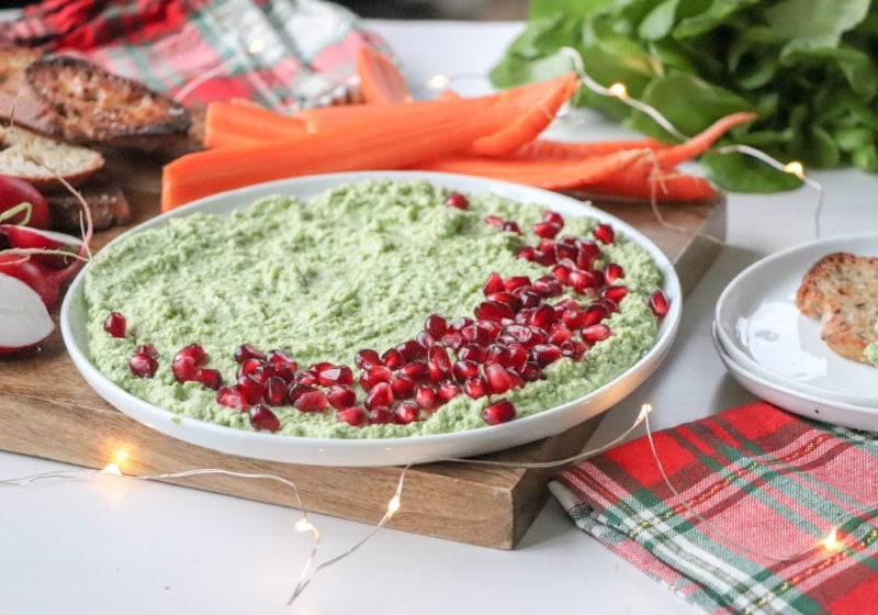 Creamy edamame dip on a white round plate placed on a wooden cutting board. Ingredients include greens, edamame, garlic, lemon, pomegranate.