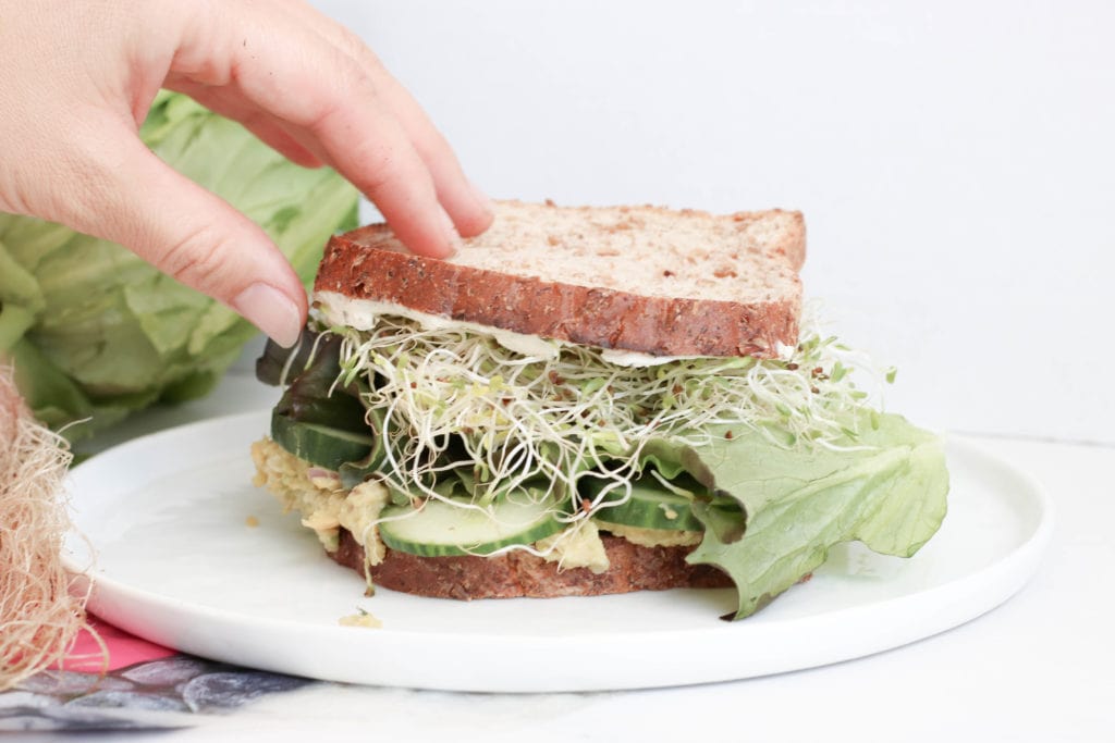 A "green goddess" sandwich on a plate with sprouts, lettuce, and cucumber seen between two whole wheat bread slices