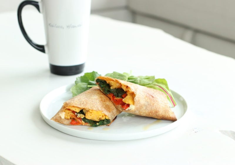A breakfast burrito cut in half and filled with eggs, spinach and peppers plated with greens on a white plate with a coffee beside it. 