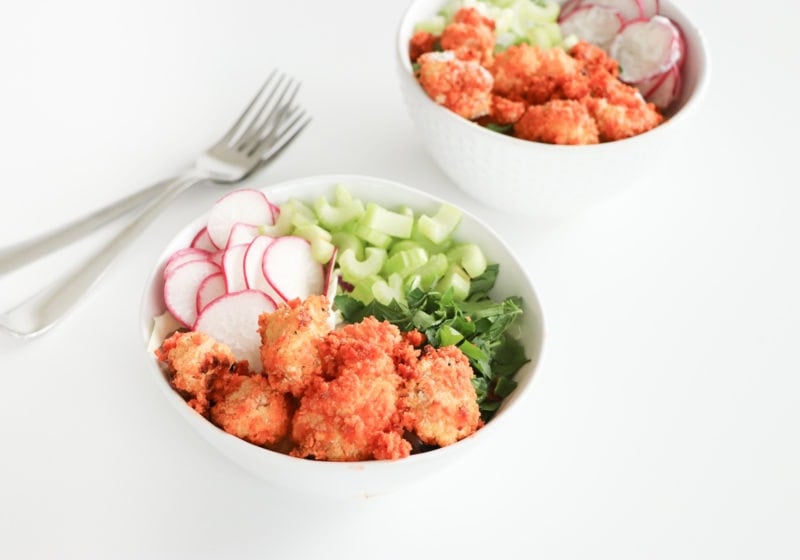 Two white bowls on a white surface with baked buffalo cauliflower, chives, celery, rice, and watermelon radish.