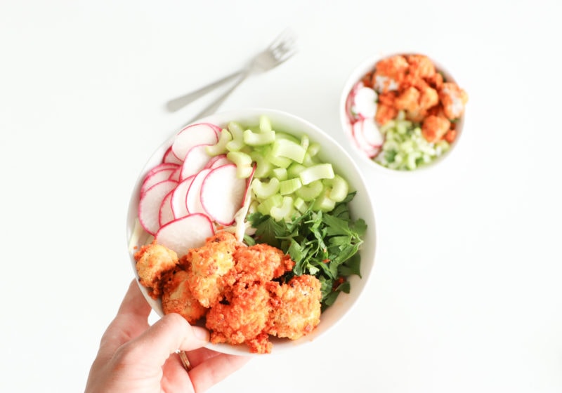 Lindsay Pleskot, RD holding a white bowl with baked buffalo cauliflower, greens, celery, and watermelon radish over a white surface.