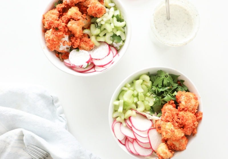 Two white bowls on a white surface with baked buffalo cauliflower, greens, and watermelon radish with a light blue kitchen towel and jar of homemade ranch dressing.