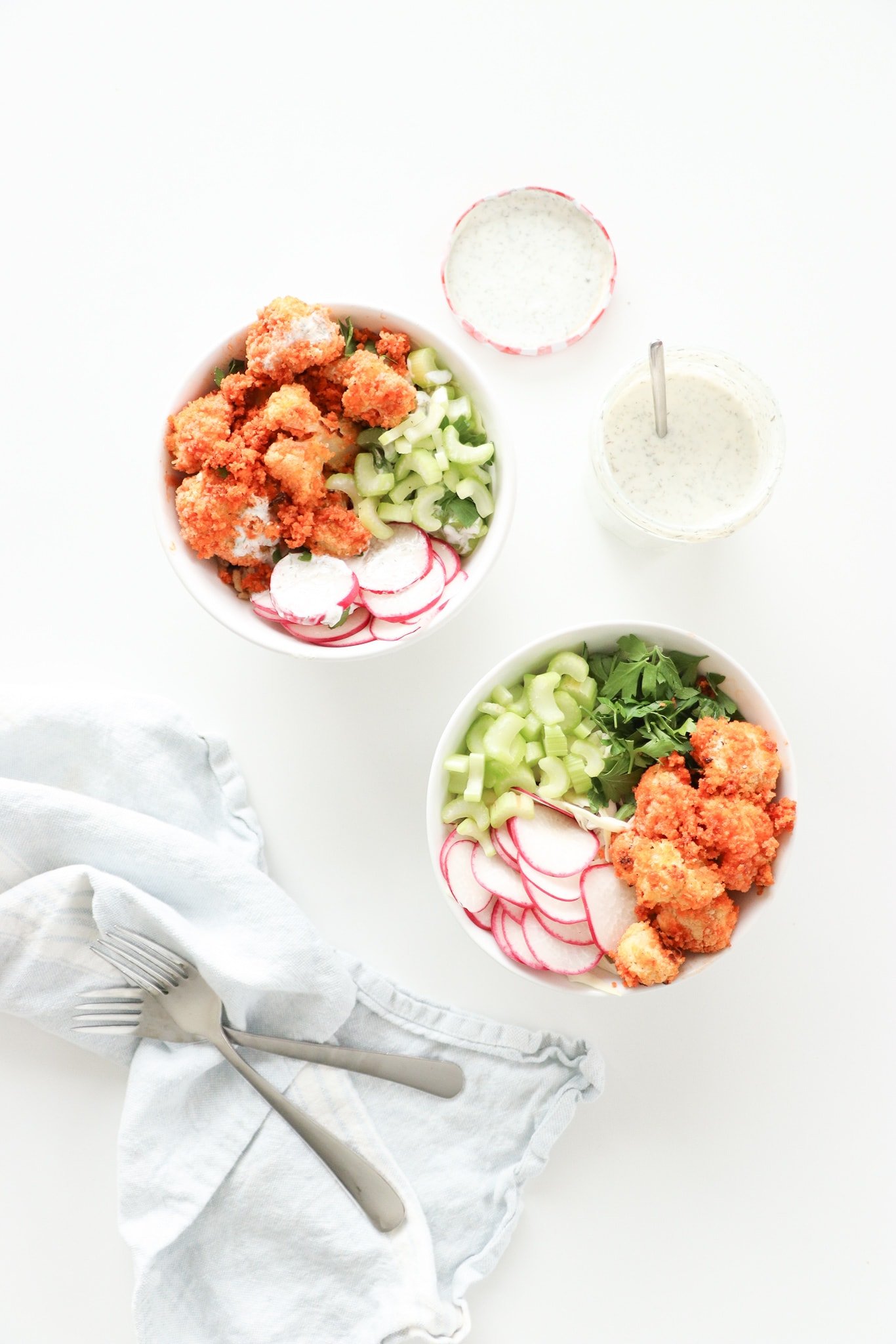 Two white bowls on a white surface with baked buffalo cauliflower, greens, and watermelon radish with a light blue kitchen towel and jar of homemade ranch dressing.