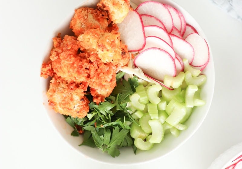A white bowl on a white surface with baked buffalo cauliflower, greens, celery, rice, and watermelon radish.