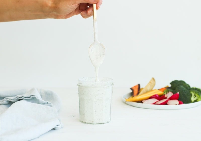 A hand holding a gold spoon drizzling greek yogurt ranch dressing from a clear jar on a white surface with veggies in the background.