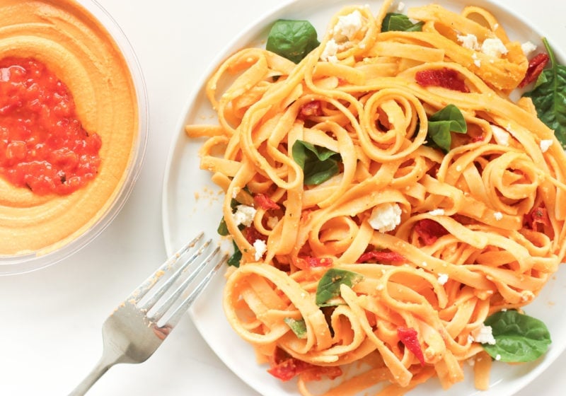 A photo of creamy red pepper pasta on a white plate. Ingredients include: fettuccine noodles, red pepper, hummust, spinach, sundried tomatoes, feta cheese.