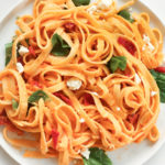 A photo of creamy red pepper pasta on a white plate. Ingredients include: fettuccine noodles, red pepper, hummust, spinach, sundried tomatoes, feta cheese.