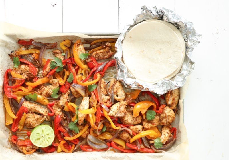 30 minute sheet pan fajitas made with simple ingredients like bell peppers, red onion, and chicken on a sheet pan over a white surface with soft shell tacos and avocados beside it.