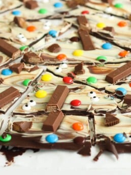 Halloween chocolate bark broken into pieces on a white surface. Ingredients include semi sweet chocolate, white chocolate, and Halloween candies.