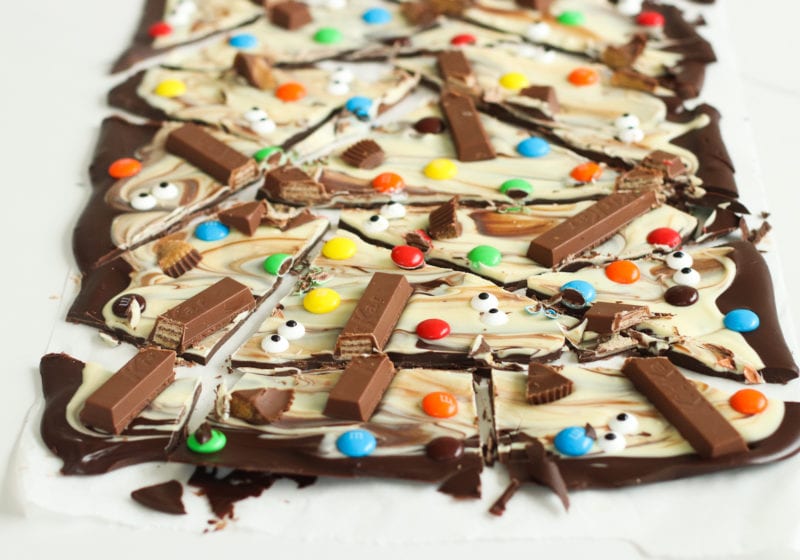 Halloween chocolate bark broken into pieces on a white surface. Ingredients include semi sweet chocolate, white chocolate, and Halloween candies.