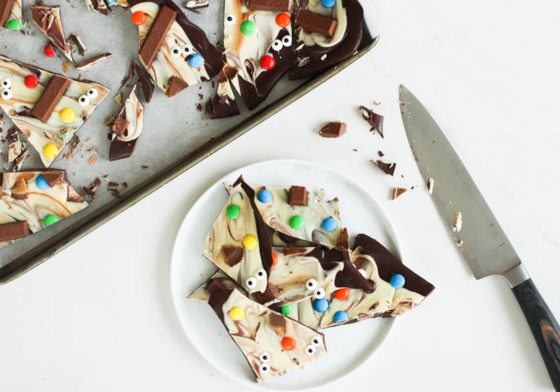 Halloween chocolate bark with semi sweet chocolate, white chocolate, halloween candies, on a baking sheet placed on a white surface with a white plate that has chopped up chocolate bark on it.