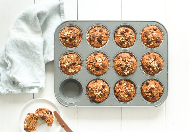 A muffin tin full of oat flour pumpkin muffins is placed on a blue napkin. On the side there is a muffin cut in half on a white plate. Recipe by lindsay pleskot registered dietitian