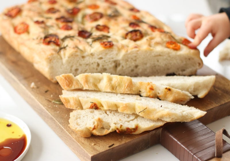 5-ingredient focaccia bread made with all purpose flour, salt, yeast, water, and olive oil topped with sundried tomatoes and rosemary placed on a wooden cutting board with a white dish beside it of balsamic vinegar. Photo features Lindsay's sons sweet hand poking the bread.