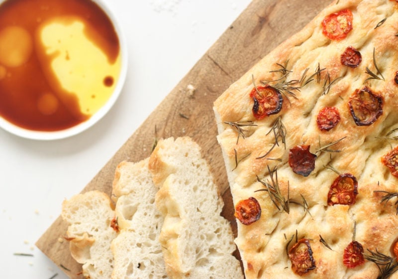 5-ingredient focaccia bread made with all purpose flour, salt, yeast, water, and olive oil topped with sundried tomatoes and rosemary placed on a wooden cutting board with a white dish beside it of balsamic vinegar.