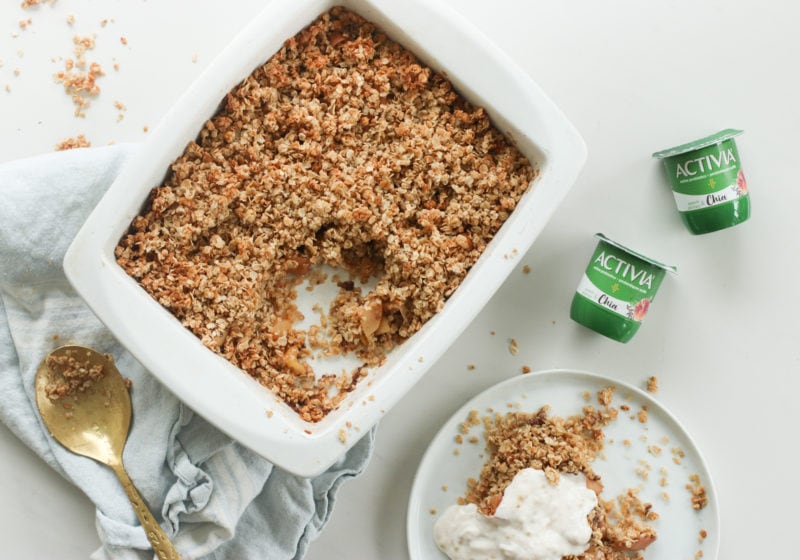 Baked breakfast apple crisp in a white dish on a white surface with Activia Chia yogurt and a kitchen towel and gold spoon beside it. A white plate is beside the dish of apple crisp with a scoop of breakfast apple crisp topped with Activia yogurt. 