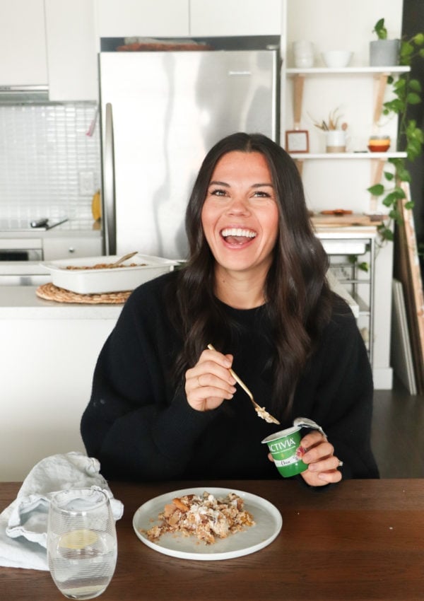 Lindsay Pleskot, Registered Dietitian wearing all black sitting at her dining room table holding Activia chia yogurt with baked apple crisp in front of her on a white plate and a light blue kitchen towel.