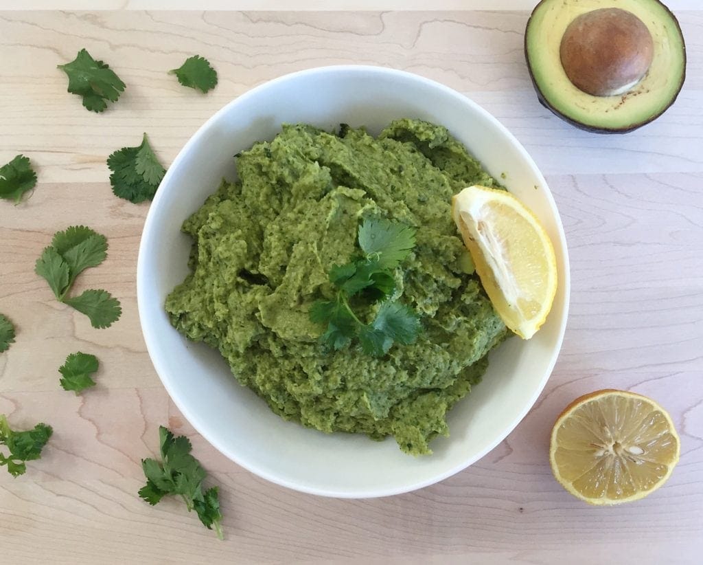Broccomole in a white bowl over a cutting board with a lemon wedge surrounded by avocado and half of a lemon. Ingredients include: broccoli, avocado, lemon juice, salt and pepper, cilantro.