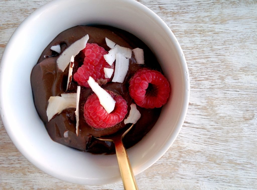 Chocolate avocado pudding in a white bowl with a gold spoon over a wood food photo board. Ingredients include: avocado, greek yogurt, cocoa powder, vanilla, honey, raspberries.