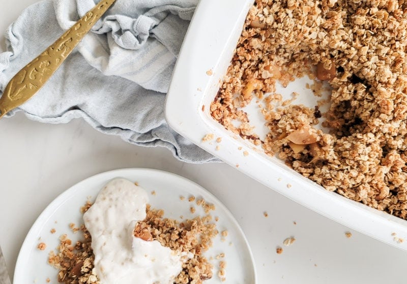 Baked breakfast apple crisp in a white baking dish served on a white plate. Ingredients include oats, almond flour, maple syrup, coconut oil, pinch of sea salt, probiotic yogurt.