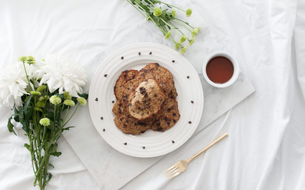 Banana oat pancakes on a white plate over a marble food board. Ingredients include banana, oats, cinnamon, chocolate chips.